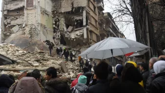 L'image montre une scène de destruction après un tremblement de terre. On peut voir un bâtiment gravement endommagé, avec des murs effondrés et des débris éparpillés. Au premier plan, une foule de personnes semble se rassembler, regardant les secours qui travaillent. Certains porteurs d'ombrelles se protègent de la pluie. L'atmosphère est sinistre, et l'on ressent une forte émotion face à la destruction causée par la catastrophe.