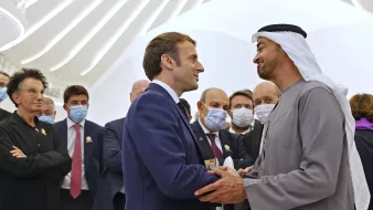 در این تصویر، یک گروه از افراد در حال گفتگو و تعامل هستند. در وسط تصویر، دو شخصیت اصلی به یکدیگر دست داده و باهم صحبت می‌کنند. یکی از آن‌ها لباس رسمی با رنگ تیره بر تن دارد و دیگری لباس سنتی اماراتی پوشیده است. بقیه افراد در پس‌زمینه نیز با لباس‌های رسمی و به نحوی متوجه این تعامل هستند. فضای تصویر روشن و مدرن به نظر می‌رسد.