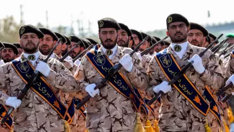 L'immagine mostra un gruppo di uomini in uniforme che sfilano. Indossano abiti mimetici e berretto, con fasce distintive sui loro uniformi. Alcuni di loro tengono in mano fucili, tutti sembrano professionali e seri. Lo sfondo è sfocato, ma suggerisce un luogo di parata o celebrazione ufficiale. L'atmosfera sembra essere di solennità e disciplina.