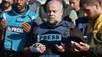 L'immagine mostra un gruppo di persone in piedi che sembrano partecipare a un momento di preghiera o riflessione. In primo piano, un uomo indossa un giubbotto con la scritta "PRESS" ed ha una fasciatura su un braccio. Gli altri membri del gruppo, che indossano abbigliamento simile, appare coinvolti nell'atto di pregare, con le mani sollevate. L'atmosfera sembra essere solenne e carica di emotività.