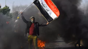 L'image montre une scène de manifestation en Irak. Un homme, portant un couvre-visage et des vêtements sombres, brandit un drapeau irakien. En arrière-plan, on peut voir des nuages de fumée noire, suggérant des feux ou des barricades sur la route. L'atmosphère semble tendue, indiquant une situation de protestation.