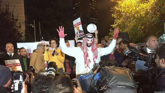 The image shows a group of people gathered for a protest or demonstration. In the foreground, a person wearing traditional attire, possibly a thobe and a headscarf, stands out. They have red paint on their hands, which could symbolize blood or a protest message. The individual is raising their hands and appears to be addressing the crowd or holding up a sign. Surrounding them are numerous cameras and reporters, indicating a media presence. The setting appears to be urban, with buildings visible in the background.