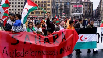 The image depicts a lively protest or demonstration taking place in an urban setting, likely in a city square. Participants are holding banners and flags, prominently featuring a design related to the Sahara. The banners include messages advocating for self-determination, with phrases like "Sahara Autodeterminazione" visible. The crowd consists of diverse individuals, including families and children, all appearing passionately engaged in the event. The background includes buildings and advertisements, adding to the urban atmosphere of the gathering. Rain can be seen drizzling, suggesting a potentially challenging weather environment for the demonstrators.