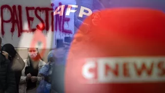 L'image présente une scène de manifestation. On peut voir un graffiti rouge avec le mot "PALESTINE" écrit en grandes lettres. En arrière-plan, on distingue des personnes rassemblées, certaines portant des masques. Des emblèmes de médias, comme "AFP" et "CNEWS", sont également visibles, suggérant une couverture médiatique de l'événement. L'atmosphère semble être chargée d'émotion et de revendication.