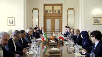L'image montre une salle de réunion avec une grande table au centre. De chaque côté de la table, plusieurs personnes sont assises, participent à une discussion. Les drapeaux de l'Iran et d'un autre pays sont visibles sur la table. L'environnement est élégant, avec des décorations raffinées et de grandes fenêtres. On peut aussi voir des fleurs au centre de la table. Les participants semblent concentrés et engagés dans la conversation.