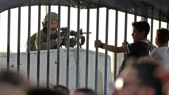 L'image montre un soldat armé stationné derrière une barrière ou un grillage. Il est en position de vigilance, regardant au loin. En face de lui, un jeune garçon semble gesticuler, tandis que d'autres enfants ou adolescents se trouvent à proximité. L'environnement semble tendu, reflétant une situation de contrôle ou de conflit. La lumière du soleil indique un moment de la journée où l'activité est encore présente.