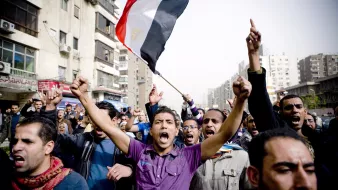 The image depicts a crowd of people in a street, likely engaged in a protest or demonstration. A man in the foreground is holding an Egyptian flag aloft and appears to be shouting passionately. The crowd around him is animated, with several individuals raising their fists and expressing strong emotions. The scene captures a sense of unity and determination among the protestors, amidst urban surroundings with buildings and visible signs of city life.