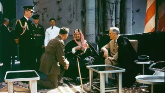 L'image montre un groupe de personnalités en discussion sur un navire. Au centre, un homme en tenue traditionnelle semble rire ou sourire, assis avec une canne. À sa gauche, un homme en habit sombre, tandis qu'un autre, en uniforme militaire, est à genoux devant eux. En arrière-plan, d'autres personnes observent la scène, dont un officier en uniforme. Le décor est richement décoré avec des tapis et des éléments nautiques.