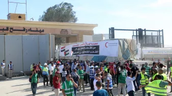 L'immagine mostra un gruppo di persone radunate davanti a un'entrata o a un confine, con bandiere nazionali visibili. Ci sono manifestanti che indossano gilet verdi e alcuni stanno tenendo striscioni. L'atmosfera sembra essere di protesta o di celebrazione, con diversi individui che interagiscono tra loro e partecipano all'evento. Sullo sfondo si vedono strutture architettoniche e una grande scritta in arabo.