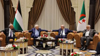 تظهر الصورة مجموعة من الأشخاص seated في غرفة مزينة بشكل أنيق، حيث يجلس أربعة رجال على أرائك مريحة. بينهم أجواء من التعاون والود، إذ يتبادلون الحديث مع قيامهم بتشابك الأيادي. في الخلفية، توجد أعلام فلسطين والجزائر، ما يشير إلى وجود موضوع يتعلق بالتعاون العربي أو القضايا السياسية بين البلدين. الطاولات أمامهم مزينة بتنسيقات من الزهور ومشروبات. الأجواء تبدو رسمية ولكن ودية في الوقت نفسه.