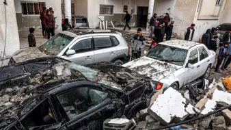 این تصویر نشان‌دهنده‌ یک صحنه ویران شده به واسطه زلزله است. چندین خودرو زیر آوارهای ساختمانی قرار گرفته‌اند و آسیب زیادی دیده‌اند. افرادی در پس‌زمینه در حال مشاهده و بررسی وضعیت هستند. آوار خانه‌ها و دیوارهای فروریخته در اطراف خودروها به چشم می‌خورد. به طور کلی، جو تصویر نشان‌دهنده اضطراب و فاجعه ناشی از زلزله است.
