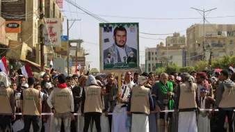 The image depicts a large crowd gathered for a rally or protest in a city, likely in Yemen. In the foreground, a group of people, some wearing beige vests, are standing behind a barricade. They are holding a large poster featuring a portrait of a man, who appears to be a political or religious leader, alongside images of other individuals. The crowd is holding flags, and buildings can be seen in the background, suggesting an urban setting. The atmosphere seems to convey a sense of solidarity or support for the figure on the poster.