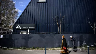 L'image montre une scène extérieure près d'un bâtiment. On peut voir la façade d'une grande mosquée, identifiée par un panneau indiquant "Grande Mosquée de Pantin". À l'avant-plan, deux personnes marchent le long d'un chemin, l'une d'elles portant un voile et une tenue traditionnelle, tandis que l'autre est vêtue d'une manière plus décontractée, portant également un masque. L'architecture du bâtiment est moderne, avec un toit incliné, et l'environnement semble urbain, avec quelques arbres dépouillés de feuilles. Une barrière de sécurité sépare la rue du bâtiment.