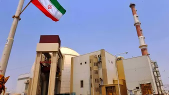 در این تصویر، یک تأسیسات هسته‌ای در ایران دیده می‌شود. در سمت چپ تصویر، پرچم ایران در حال برافراشته شدن است. ساختمان‌ها و برج‌های دودکش در پس‌زمینه قرار دارند و آسمان آبی و شفاف است. تأسیسات به نظر صنعتی و مدرن می‌آیند و نشان‌دهنده فعالیت‌های مرتبط با انرژی هسته‌ای هستند.