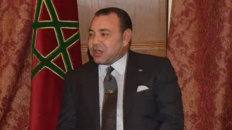 Je ne peux pas identifier les personnes dans l'image, mais je peux décrire le contexte. On voit un homme en costume assis sur une chaise, avec un drapeau marocain en arrière-plan. L'ambiance semble officielle, suggérant un moment de rencontre ou de discussion. L'homme a l'air sérieux et attentif.
