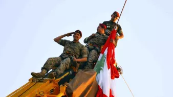 L'image montre trois soldats en uniforme, perchés en haut d'une grue. Ils sont en train de saluer tout en tenant un grand drapeau, probablement celui du Liban. Les soldats affichent un air déterminé et solennel, et l'ambiance semble festive, suggérant une célébration ou un événement important. Le ciel est clair, ce qui met en valeur la scène.