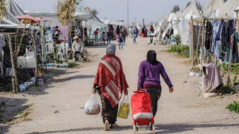L'image montre un camp de réfugiés ou un espace similaire, avec plusieurs tentes dressées de chaque côté d'un chemin central. Deux femmes marchent le long de ce chemin, portant des sacs et un chariot. L'une d'elles porte une couverture ou un châle coloré. En arrière-plan, on peut apercevoir d'autres personnes et des tentes, suggérant une atmosphère de communauté. Le paysage semble rural et ensoleillé, avec des éléments de végétation autour.