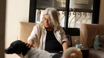 Nell'immagine, vediamo una donna anziana con capelli bianchi e lunghi che indossa occhiali e una camicia chiara. È seduta su un divano in una stanza luminosa, con una finestra sullo sfondo che lascia entrare la luce. Accanto a lei, un cane bianco con macchie nere si avvicina, mentre la donna sembra accarezzarlo con affetto. L'atmosfera è calda e accogliente, riflettendo un momento di intimità tra la donna e il suo animale domestico.
