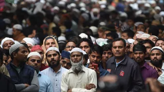 La imagen muestra una multitud de personas, muchas de las cuales llevan puestos kufis o tapones. La mayoría de las personas parecen estar prestando atención a algo en frente de ellos, algunos lucen serios y concentrados. Hay una gran diversidad de edades entre los asistentes y la atmósfera parece de expectativa. El fondo está lleno de más personas, lo que indica que es una reunión o evento masivo.