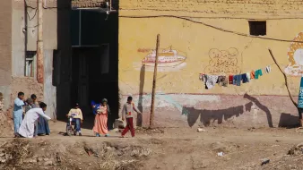 The image depicts a scene from a rural or semi-urban area, featuring a group of people interacting outside. A child in a wheelchair is being pushed forward by an adult, while two other children walk beside them. In the background, there is a building with a yellow wall that has some painted decorations or text. Above this, a clothesline displays various articles of clothing hanging to dry. The ground appears uneven, with some debris scattered around. Overall, the scene conveys everyday life in a community setting.