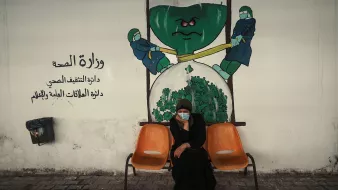 الصورة تظهر امرأة جالسة على كرسيين في منطقة تبدو وكأنها مركز صحي أو مستشفى. تحمل المرأة ملامح الجدية، وتضع كمامة على وجهها. خلفها يوجد جدار م painted يوضح رسماً يجسد فكرة وزارة الصحة، حيث يظهر شخصيتان ترتديان بزة وقاية، تسعيان لشد أو دفع شيء كبير يشبه الخضار أو الملفوف. الألوان المستخدمة في الجدار تعكس موضوع الصحة، مع تركيبات واضحة ورموز مثل القفازات والكمامات.