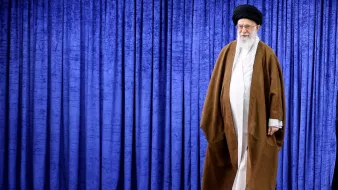 در تصویر، فردی با لباس سنتی روحانی به رنگ‌های قهوه‌ای و سفید دیده می‌شود. او کلاهی مشکی بر سر دارد و ریش بلندی دارد. پس‌زمینه تصویر شامل پرده‌ای به رنگ آبی است. فرد در حال حرکت به سمت سمت راست تصویر قرار دارد. این تصویر به طور کلی حس وقار و احترام را القا می‌کند.