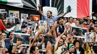 تظهر الصورة حشودًا كبيرة من الناس في تجمع أو مظاهرة. يبدو أنهم يحملون لافتات وصورًا، ويظهر في المقدمة شخص يرفع يده ويبدو أنه يتحدث إلى الجمهور. الناس في الخلف يحملون أعلامًا وصورًا، ما يدل على أنهم يعبرون عن مشاعر قوية، ربما تتعلق بقضية سياسية أو اجتماعية. يبدو أن الجو مليء بالحماس والتفاعل.