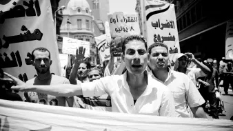 L'image montre une manifestation animée en noir et blanc. Des participants tiennent des pancartes et expriment leur mécontentement. Au premier plan, un homme semble parler avec passion, tandis que d'autres personnes en arrière-plan lèvent les bras et crient. Les banderoles sont en arabe, indiquant des revendications sociales ou politiques. L'ambiance est dynamique et engagée.