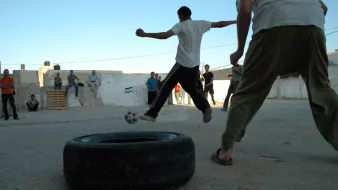 L'immagine mostra un gruppo di persone che si stanno divertendo in un ambiente all'aperto. Alcuni ragazzi stanno giocando a calcio, utilizzando un pneumatico come obiettivo. Si vedono diverse persone in piedi attorno, probabilmente che osservano la partita. La scena si svolge su un terreno sterrato, con muri bianchi sullo sfondo e un cielo azzurro. L'atmosfera è di divertimento e convivialità.