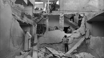 L'image montre un bâtiment en ruines, visiblement endommagé par un conflit ou un désastre. Les murs sont effondrés et des débris jonchent le sol. Plusieurs personnes se trouvent dans l'espace, certains semblent fouiller parmi les débris, tandis que d'autres observent la scène depuis un étage supérieur. L'ambiance est sombre et témoigne des conséquences dévastatrices sur la vie humaine et les infrastructures.