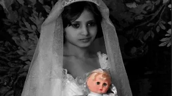 L'image montre une jeune fille portant une robe blanche et un voile. Elle tient une poupée dans ses bras. Le fond est sombre et texturé, ce qui met en valeur l'apparence délicate de l'enfant et de sa tenue. L'expression de son visage semble mélancolique et contemplative.