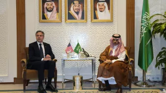 L'image montre deux hommes assis côte à côte dans un cadre formel. À l'arrière-plan, il y a trois portraits encadrés de dirigeants saoudiens. Sur une table entre eux, se trouvent des éléments décoratifs. L'un des hommes porte un costume sombre et l'autre est habillé en tenue traditionnelle saoudienne. À droite, on aperçoit un drapeau saoudien. L'atmosphère semble officielle et diplomatique.