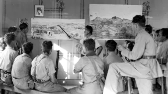 L'image montre un groupe de personnes assises sur des bancs, attentives à un homme qui semble donner une présentation devant des tableaux accrochés au mur. Les tableaux illustrent des paysages, l'un représentant probablement un village ou une scène de la nature. L'environnement est dépouillé, avec des murs simples et une lumière naturelle qui pénètre à travers une fenêtre. Les participants, majoritairement en vêtements uniformes, semblent captivés par l'explication de l'intervenant. Il y a aussi un portrait encadré sur le mur, ajoutant un élément de contexte à la scène.