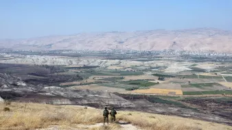 در این تصویر، دو نفر در حال ایستادن بر روی یک تپه یا ارتفاع هستند و به منظرۀ وسیع و زیبای زیر پای خود نگاه می‌کنند. در زیر، دشت‌ها و زمین‌های کشاورزی با رنگ‌های مختلف دیده می‌شوند. دامنه‌های کوه‌ها در دوردست قرار دارند و آسمان آبی و صاف به چشم می‌خورد. فضا به نظر آرام و دلنشین می‌رسد و نشان‌دهندهٔ مناظر طبیعی و انسانی در منطقه‌ای کوهستانی است.