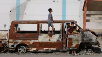 L'image montre un groupe d'enfants jouant autour d'une vieille fourgonnette rouillée et endommagée. Un enfant est debout sur le toit de la voiture, tandis que d'autres sont assis et debout autour d'elle, semblant s'amuser malgré l'environnement délabré. En arrière-plan, on aperçoit un bâtiment aux murs abîmés. L'atmosphère semble à la fois joyeuse et nostalgique, illustrant la résilience des enfants dans un cadre difficile.