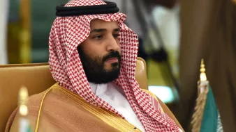 L'image montre un homme portant un long vêtement traditionnel arabe, composé d'une robe beige et d'un headdress appelé "ghutrah" ou "keffieh", qui est noué avec un bandeau noir. Il a une expression sérieuse et se trouve assis sur une chaise. En arrière-plan, des drapeaux peuvent être aperçus, suggérant un événement officiel ou une rencontre.