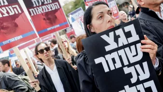 Nell'immagine si vede una manifestazione di persone vestite con abiti neri, probabilmente avvocati, che tengono cartelli. In primo piano, una donna tiene un grande cartello con un messaggio ben visibile. Sullo sfondo, ci sono altre persone con cartelli, in un'atmosfera di protesta. Le persone sembrano serie e impegnate nella loro causa. L'ambiente è quello di una manifestazione pubblica, con molta partecipazione.