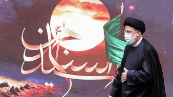 در این تصویر، یک مرد در لباس رسمی و با عمامه مشکی در حال راه رفتن است. او ماسک به صورت دارد و پشت او تصاویری از یک پس‌زمینه هنری با رنگ‌های گرم و نمادهایی مربوط به تاریخ یا فرهنگ دیده می‌شود، از جمله پرچم سبز. متن خوشنویسی شده‌ای در تصویر وجود دارد که به زبان فارسی نوشته شده است. این تصویر ممکن است به یک مراسم مذهبی یا ملی مرتبط باشد.