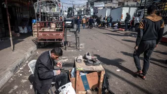 در تصویر، مردی در حال تعمیر یا واکس زدن کفش‌ها است. او روی یک صندلی نشسته و وسایل کارش را بر روی یک میز کوچک چوبی قرار داده است. در اطراف او، افرادی در حال عبور و مرور هستند و تعدادی خودروی باربری در پس‌زمینه دیده می‌شود. فضایی شلوغ و شهری به نظر می‌رسد و فعالیت‌های روزمره در حال انجام است. چند کشیده و بقیه اجناس نیز در اطراف قرار دارند.