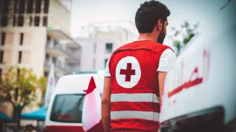 L'image montre un homme portant une veste rouge avec une croix blanche, typique des membres d'une organisation de secours, comme la Croix-Rouge. Il se tient de dos, observant une scène à proximité de véhicules de secours. En arrière-plan, on aperçoit des bâtiments modernes et des drapeaux, ce qui pourrait indiquer un environnement urbain. L'atmosphère semble calme, mais le contexte suggère une implication dans des missions sanitaires ou d'assistance.