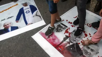 در این تصویر، تعدادی از افراد با پا بر روی پوسترهایی که عکس‌هایی از شخصیت‌های سیاسی معروف را نشان می‌دهند، ایستاده‌اند. یکی از پوسترها به تصویر نتانیاهو مربوط می‌شود و دیگری شامل تصاویری از ترامپ و یک شخصیت دیگر است. نشان‌های خون و نوشته‌ها بر روی پوسترها به وضوح دیده می‌شوند و این تصویر به نظر می‌رسد در یک تظاهرات یا اعتراض گرفته شده باشد.