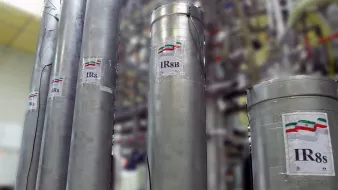 تظهر في الصورة مجموعة من الأسطوانات المعدنية، وكل منها يحمل ملصق مكتوب عليه "IR8" أو "IR8s" مع رمز علم دولة إيران. خلف هذه الأسطوانات، يمكن رؤية مجموعة من الأنابيب والتجهيزات الأخرى، مما يشير إلى أنها قد تكون جزءًا من منشأة صناعية أو مركز بحث علمي. الجو العام في الصورة يعكس بيئة تقنية عالية ومجهزة بأجهزة متطورة.