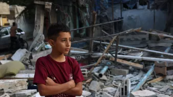 L'image montre un jeune garçon debout, les bras croisés, au milieu des débris d'un bâtiment effondré. Il semble pensif et préoccupé, exprimant une certaine gravité face à la destruction qui l'entoure. Les morceaux de béton et les matériaux épars suggèrent un environnement post-catastrophe, tandis que l'arrière-plan laisse entrevoir d'autres structures endommagées. L'atmosphère générale est sombre et témoigne d'une situation difficile.