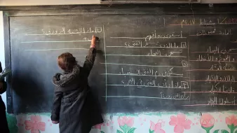 الصورة تظهر فتاة صغيرة تقف أمام سبورة سوداء في فصل دراسي. هي تكتب على السبورة، والسبورة تحتوي على قائمة مكتوبة بخطوط مختلفة. الجدران مزينة برسومات زهور ملونة، مما يضفي جواً من الحيوية على المكان. الفتاة ترتدي معطفاً واقفاً، وتبدو مت concentrating في ما تفعله.