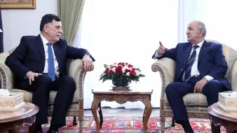 L'image montre deux hommes assis dans un environnement décoré de façon formelle. Ils sont en train de discuter, chacun semblant attentif à l'autre. Le premier homme, habillé en costume avec une cravate, se tourne légèrement vers le second. Ce dernier, également en costume, gesticule avec une main, ce qui pourrait indiquer qu'il est en train de faire un point ou de partager une idée. Entre eux, il y a une table basse ornée d'un arrangement floral. L'arrière-plan est lumineux et dégage une ambiance de rencontre officielle. Des éléments décoratifs, comme des rideaux et un tapis, ajoutent une touche élégante à la scène.