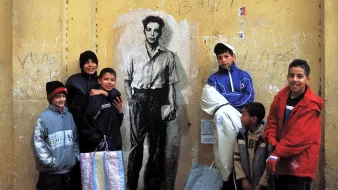 The image features a group of children standing in front of a large mural on a yellow wall. The mural depicts a black-and-white portrait of a young man in a shirt. The children are dressed in casual, colorful clothing, with some wearing hats. They appear to be interacting playfully, with a sense of camaraderie among them. The background shows some graffiti and markings, adding to the urban setting.
