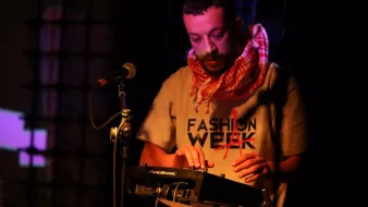 L'image montre un homme en train de performer sur scène. Il porte un t-shirt avec le mot "FASHION" écrit dessus et un foulard autour du cou. Il utilise un petit appareil électronique, probablement un synthétiseur ou un contrôleur musical. Le fond est légèrement éclairé avec des lumières colorées, créant une ambiance dynamique. Le microphone est également visible à côté de lui, indiquant qu'il pourrait chanter ou parler en même temps. L'atmosphère semble électrique et captivante.