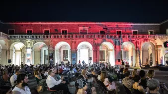 L'immagine mostra una grande sala all'aperto, con archi eleganti che circondano lo spazio. È sera e l'ambiente è illuminato da luci soffuse, creando un'atmosfera suggestiva. In primo piano, un folto pubblico è seduto su sedie disposte in file, mentre alcuni spettatori osservano attentamente. La scena trasmette un senso di partecipazione a un evento culturale o a una performance artistica. I dettagli architettonici dell'ambiente circostante sono ben visibili, contribuendo all'eleganza della location.