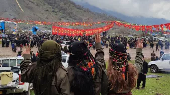 L'image montre un groupe de trois personnes vues de dos, faisant un signe de paix avec leurs mains. Elles portent des vêtements traditionnels et des foulards colorés. En arrière-plan, on peut voir une foule rassemblée pour un événement, avec des banderoles rouges décorées de symboles. Le paysage environnant est montagnard et sous un ciel nuageux. L'atmosphère semble festive et solennelle.