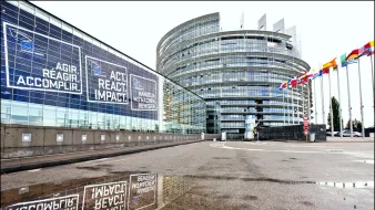 L'image représente un bâtiment moderne avec une architecture distinctive, probablement un siège d'institution européenne. On peut voir des fenêtres en verre réfléchissant des phrases comme "AGIR, RÉAGIR, ACCOMPLIR" en plusieurs langues. À droite, il y a une série de drapeaux représentant différents pays, tandis qu'une flaque d'eau au premier plan reflète l'édifice et les messages affichés sur la façade. L'ensemble donne une impression de dynamisme et de modernité.